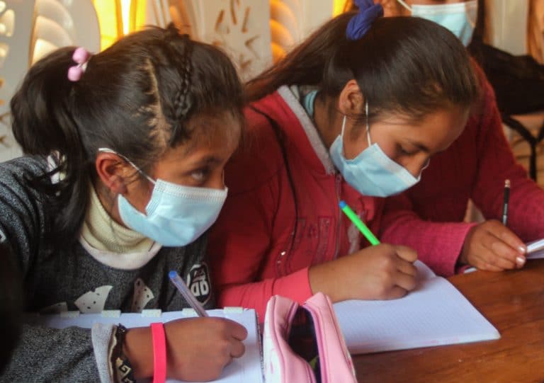 Two girls sitting at a desk working on schoolwork; the one on the left is looking up