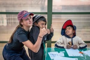 Volunteer in a cap smiling next to a boy giving a thumbs up in a classroom