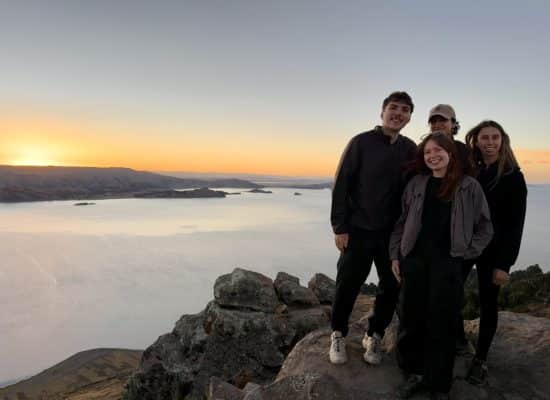 Four Globalteer volunteers smiling on a rocky viewpoint above Lake Titicaca at sunset"